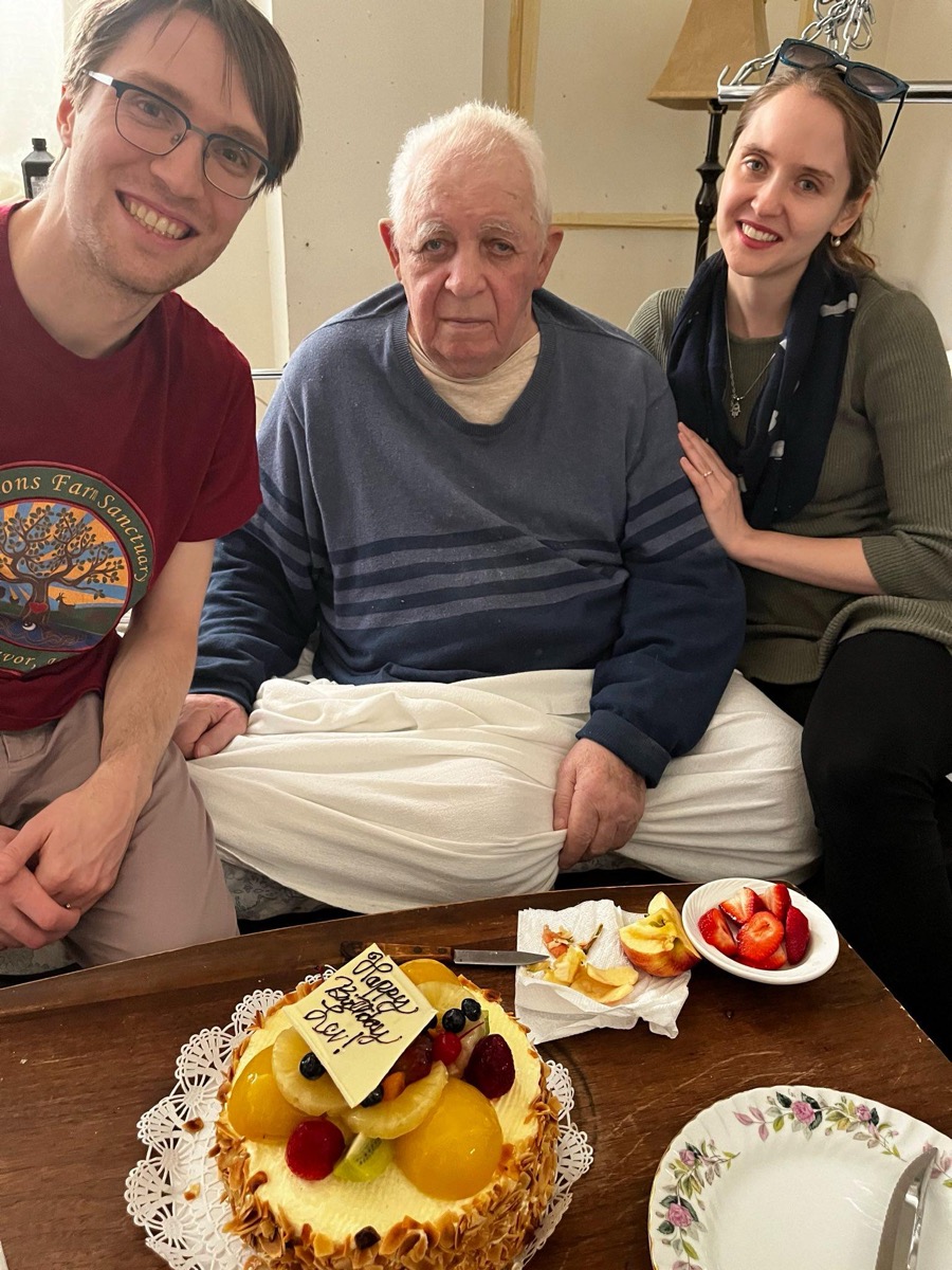 Smiling man (me) and woman (marina) on the left and right of an elderly man (Lev), with a cake in front of him reading "Happy Birthday Lev"